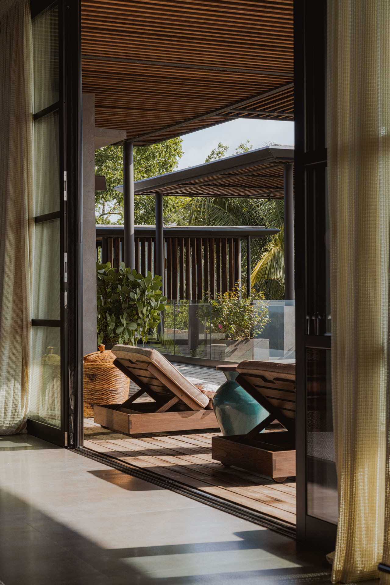 View through open sliding glass doors onto a wooden terrace with lounge chairs, sheltered by a deep slatted timber overhang at Dos Vistas in Cabuya, Costa Rica designed by Studio Saxe.