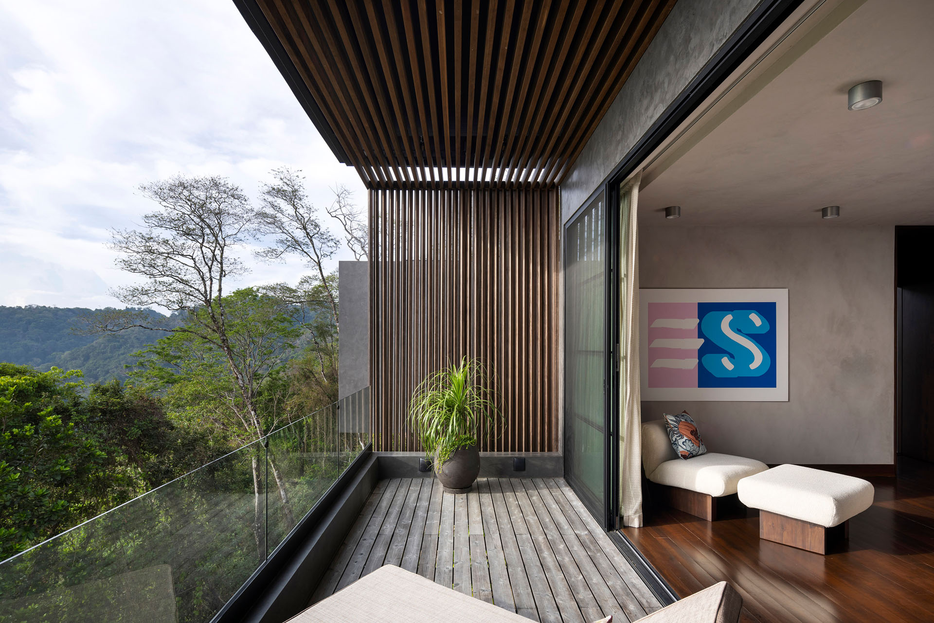 Private wooden balcony featuring a glass railing, deep timber overhang, and vertical wooden louvers connected to an interior bedroom overlooking the jungle at Dos Vistas in Cabuya, Costa Rica designed by Studio Saxe.