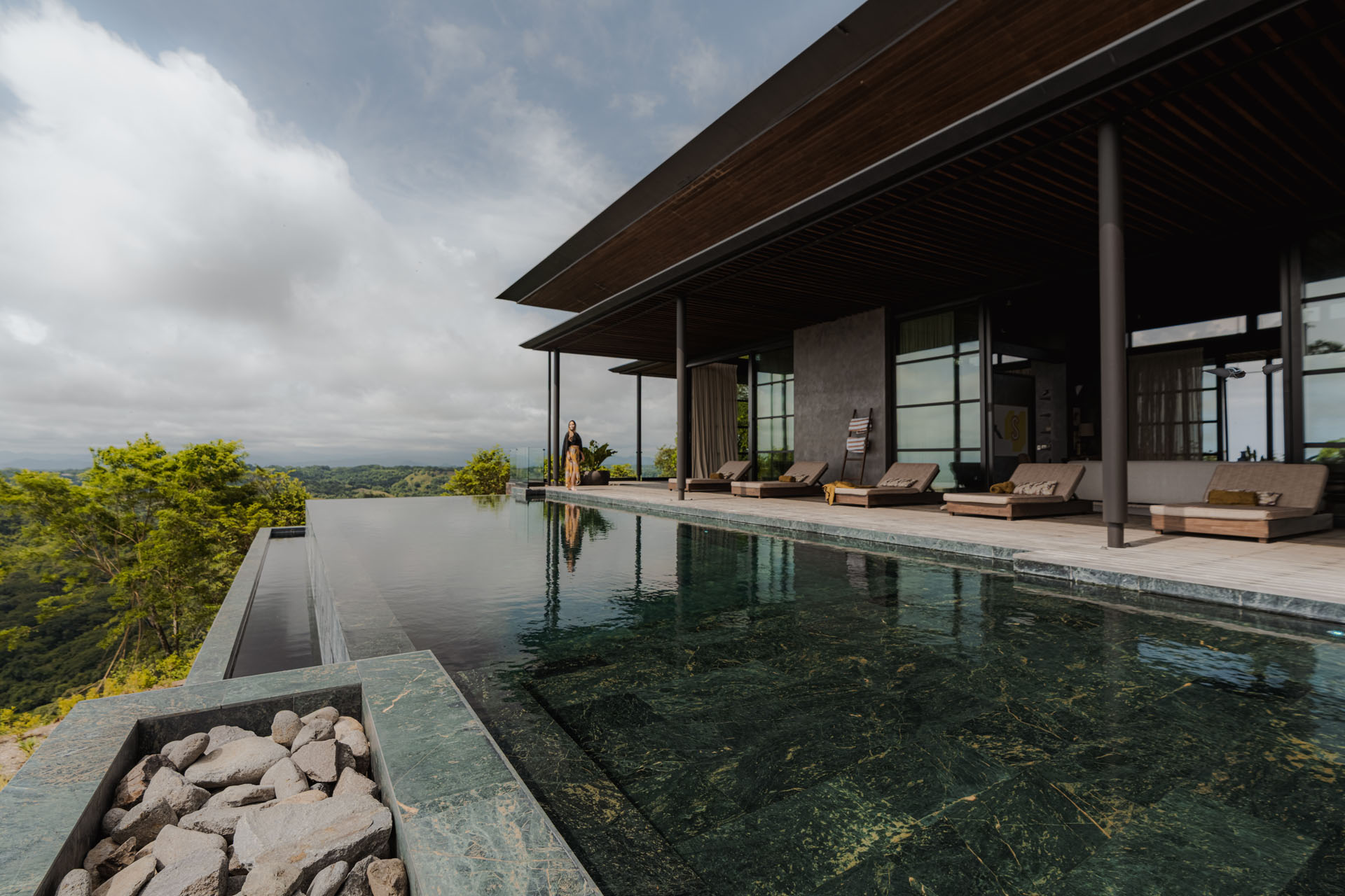 Green stone infinity pool and wooden terrace with lounge chairs beneath a large timber roof overhang at Dos Vistas in Cabuya, Costa Rica by Studio Saxe.