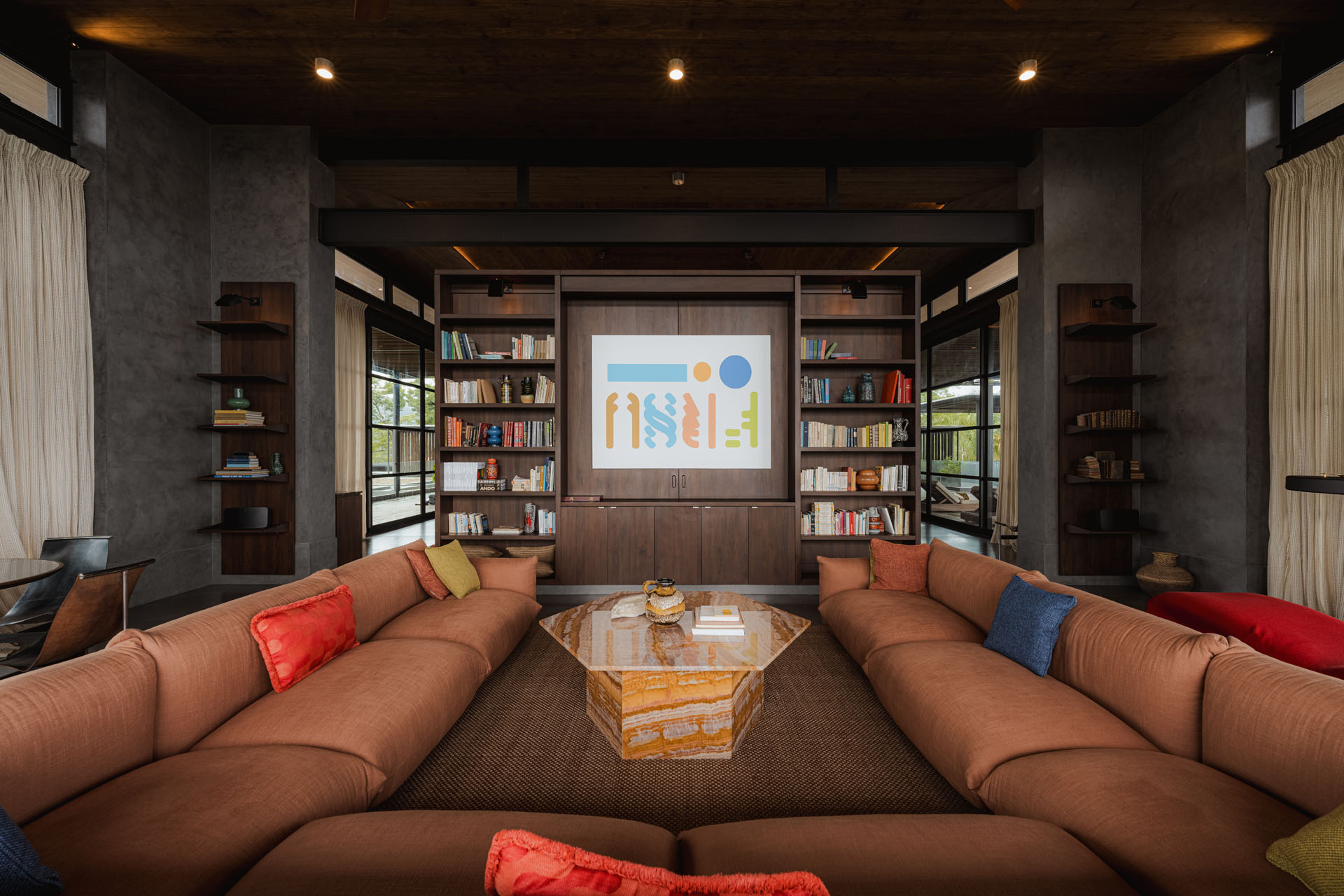 Symmetrical interior view of a large sunken lounge space featuring a U-shaped brown sofa, custom wooden bookshelves, and an octagonal stone coffee table at Dos Vistas in Cabuya, Costa Rica by Studio Saxe.