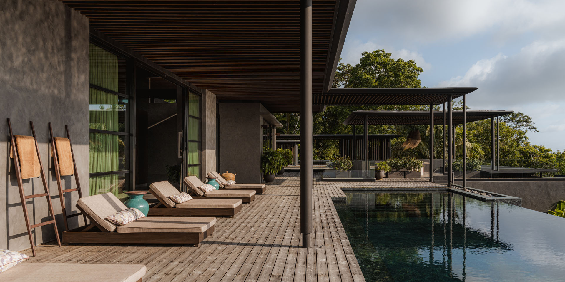 Shaded wooden pool terrace with lounge chairs next to a dark infinity pool beneath a deep timber roof overhang at Dos Vistas in Cabuya, Costa Rica by Studio Saxe.