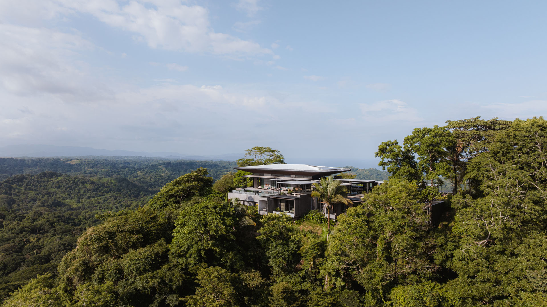 Wide aerial view of the Dos Vistas residence situated on a mountain ridge overlooking the dense tropical jungle and distant ocean in Cabuya, Costa Rica by Studio Saxe.