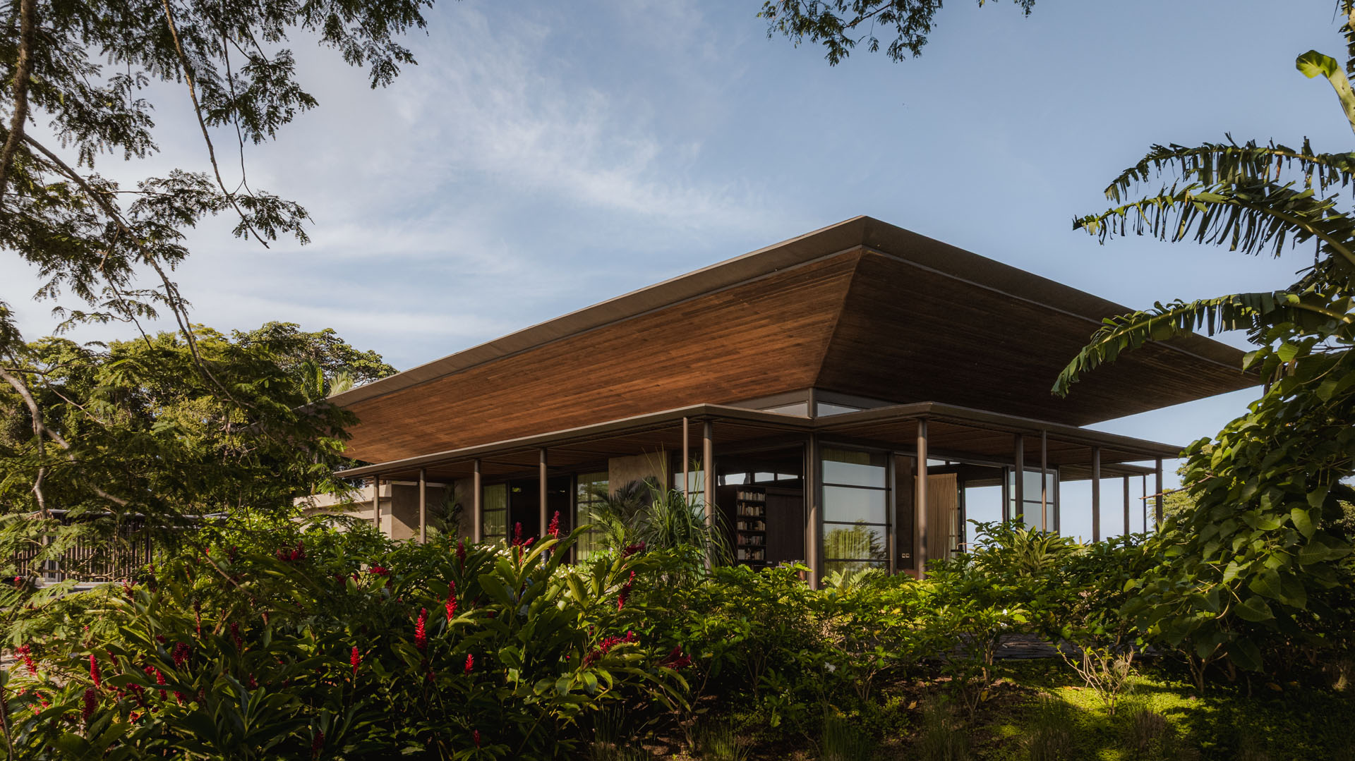 Exterior view of the main glass living pavilion and large timber roof surrounded by vibrant tropical gardens and red ginger plants at Dos Vistas in Cabuya, Costa Rica by Studio Saxe.