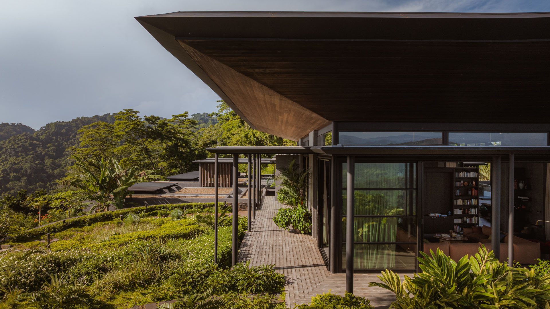 Exterior view along the shaded wooden terrace, looking past steel columns and sliding glass doors toward the planted roofs at Dos Vistas in Cabuya, Costa Rica by Studio Saxe.
