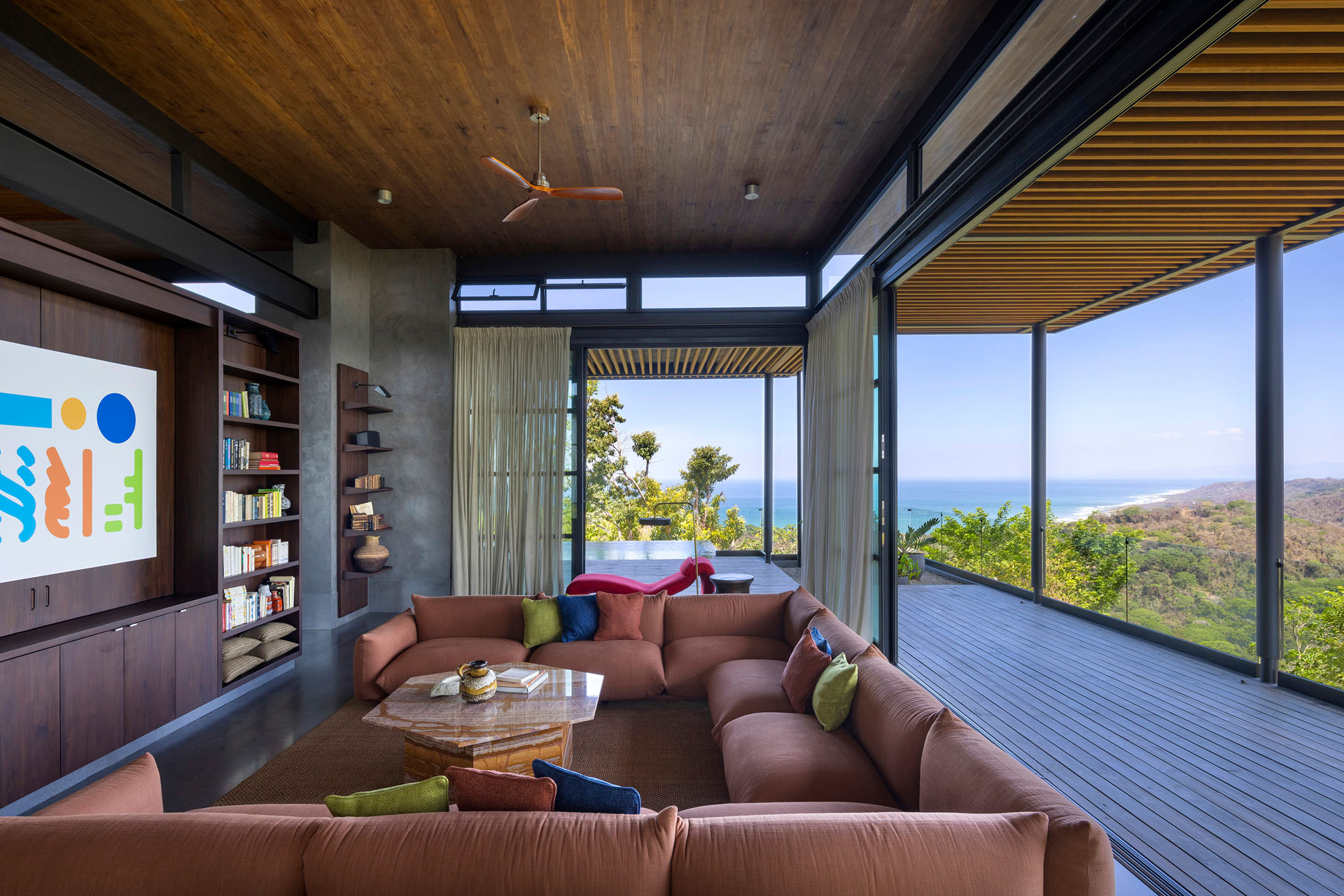 Interior living room with exposed concrete walls, a large corner sofa, and floor-to-ceiling glass sliding doors opening to expansive ocean views at Dos Vistas in Cabuya, Costa Rica by Studio Saxe.