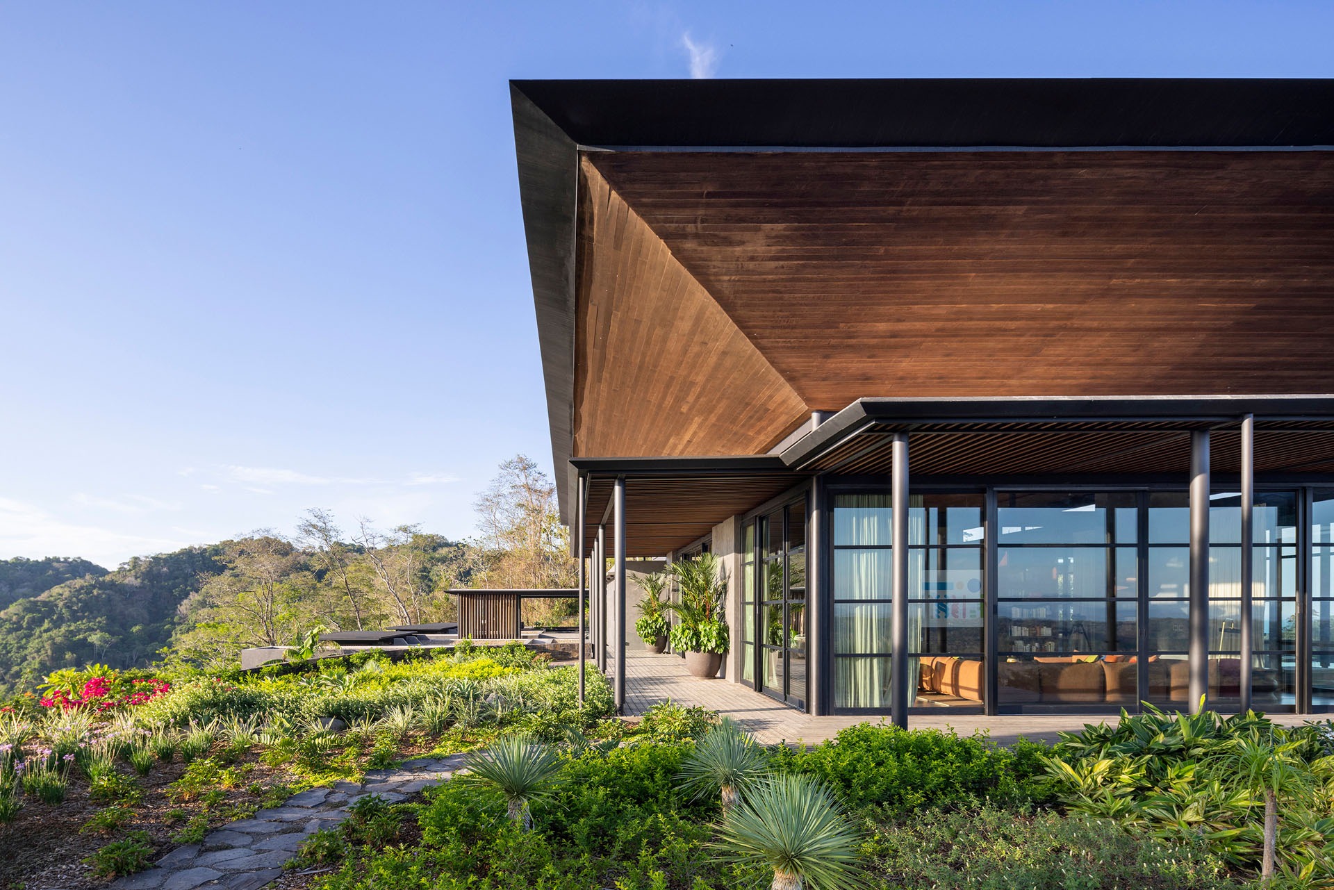 Exterior view of the main living pavilion featuring a large overhanging timber roof and floor-to-ceiling glass walls surrounded by tropical gardens at Dos Vistas in Cabuya, Costa Rica by Studio Saxe.