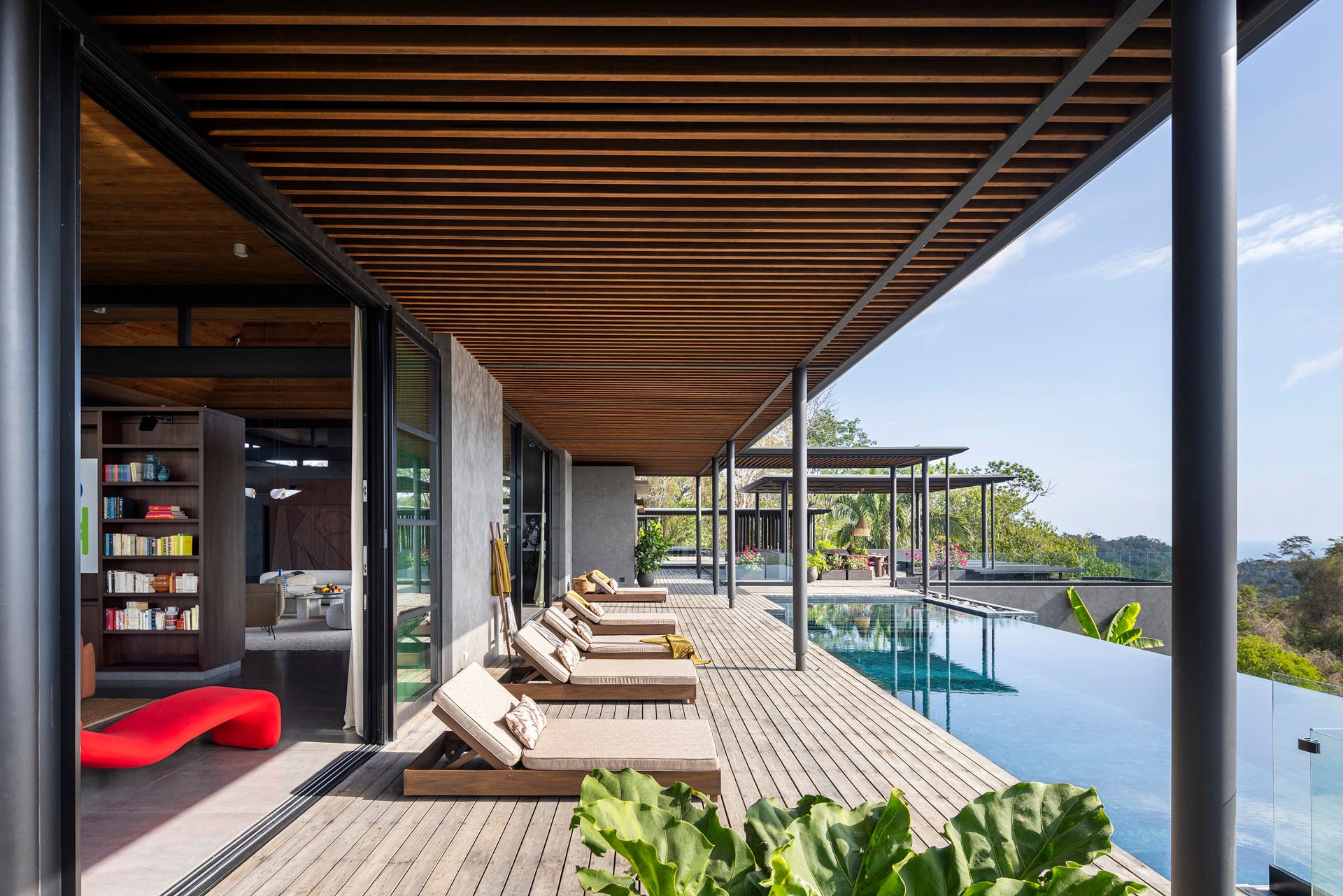 Wooden outdoor terrace with an infinity pool and lounge chairs beneath a deep slatted timber overhang at Dos Vistas in Cabuya, Costa Rica designed by Studio Saxe.